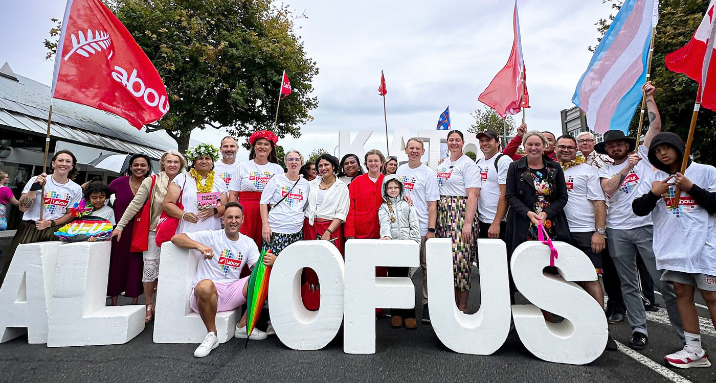 Labour MPs including Chris Hipkins, Shanan Halbert, Carmel Sepuloni, and Willow-Jean Prime standing in front of the 'All of us' signage at the Auckland Pride event in 2025, wearing the Labour Pride t-shirts designed by Bradley Pratt.